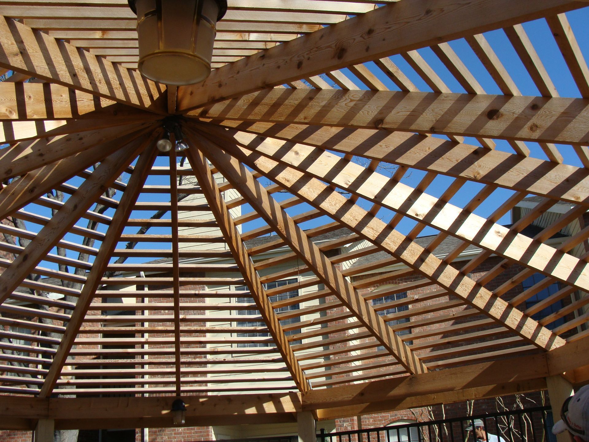 Wooden pergola roof with a radiating, geometric slat design, viewed from underneath against a bright blue sky.