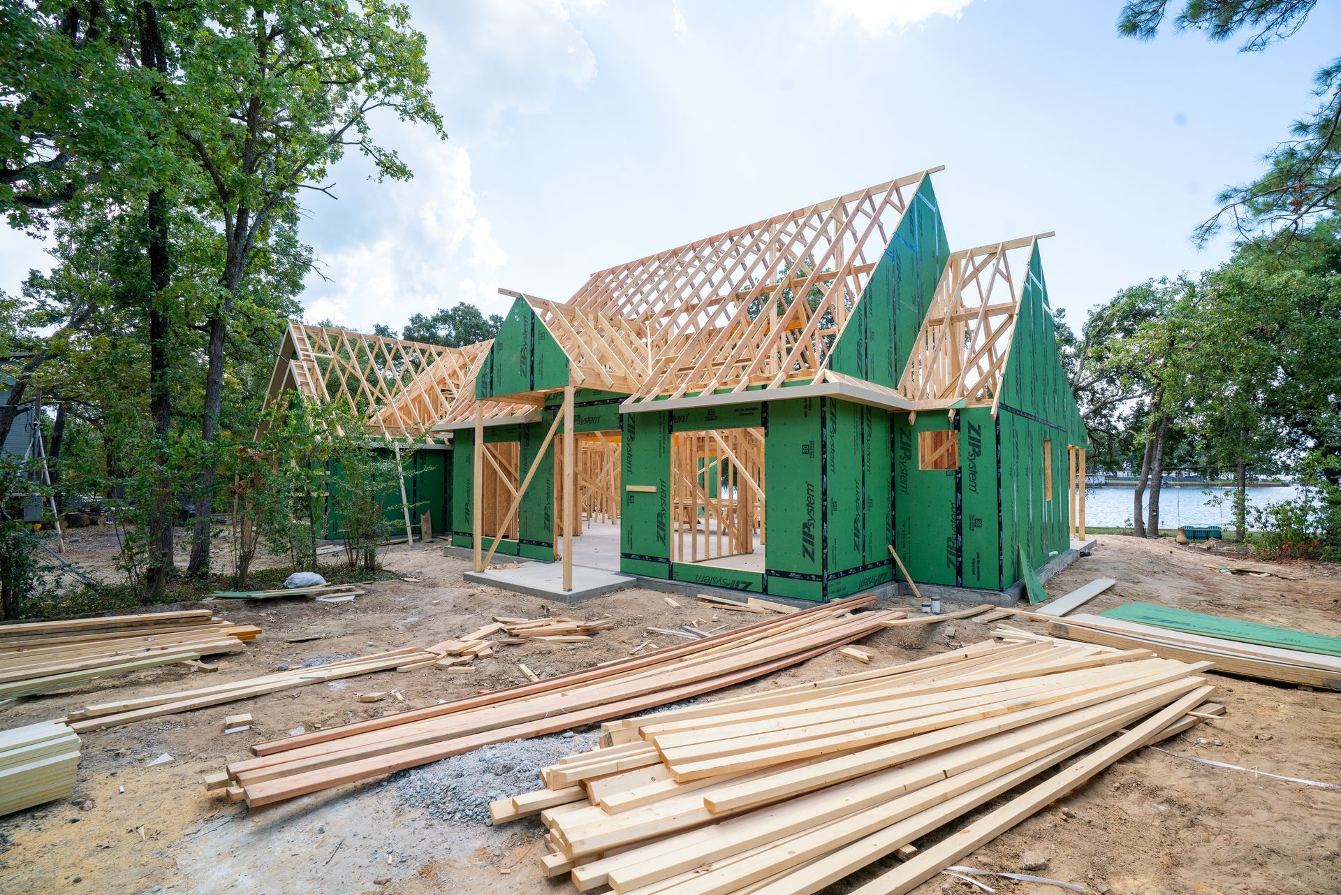 A house under construction with a wooden frame and green exterior panels, surrounded by trees and stacked lumber.