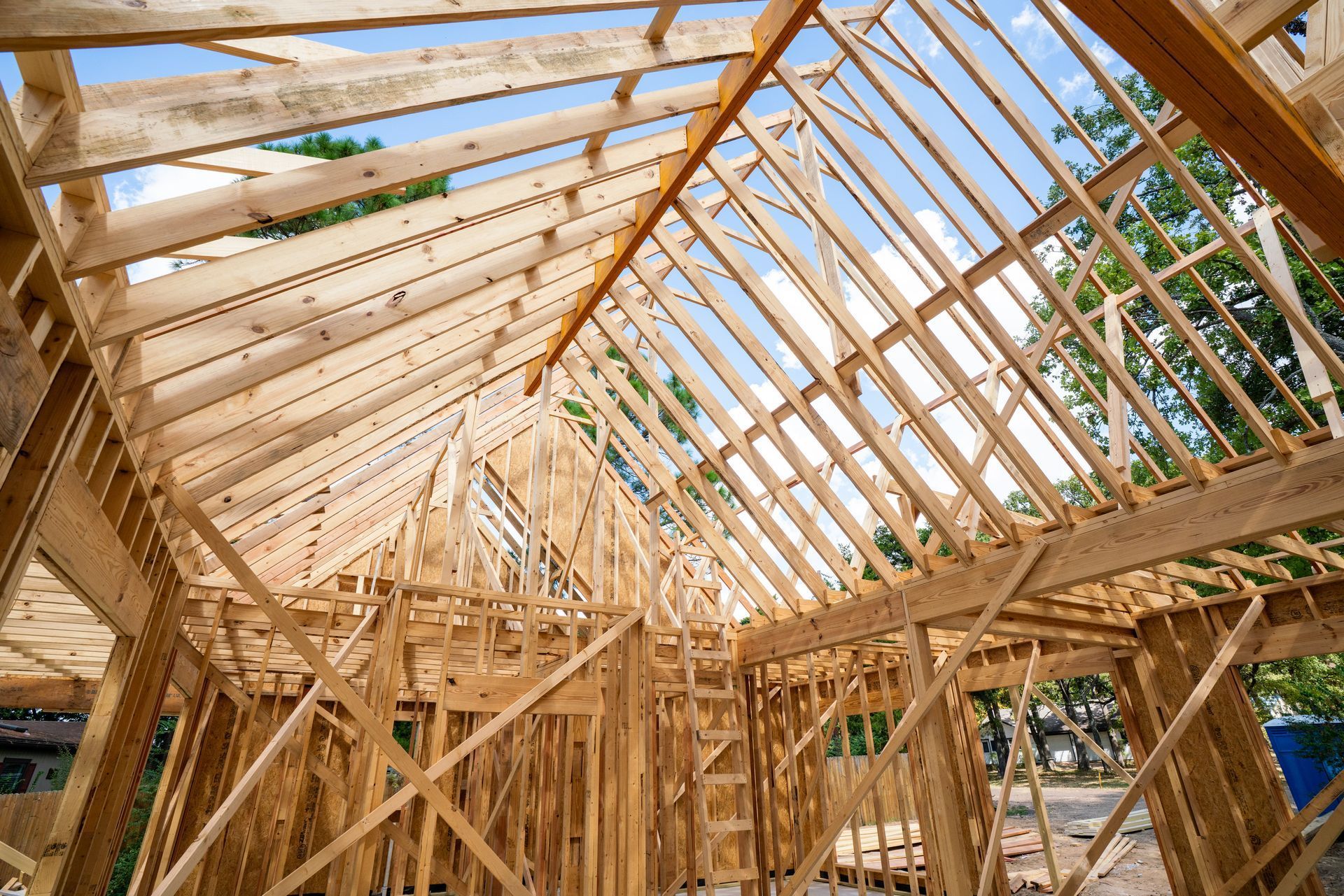 The wooden frame of an unfinished house under construction, showing rafters and roof supports against a blue sky.