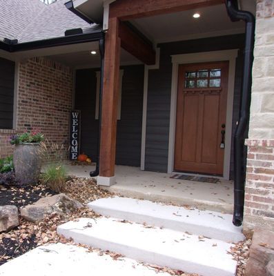 A wooden front door under a covered porch with a stone planter, a welcome sign, and concrete steps leading to the entrance.