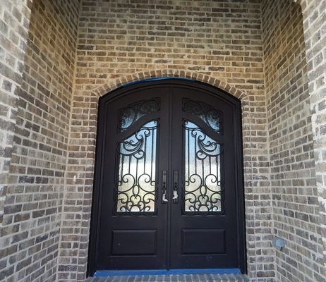 A dark double door with decorative iron grilles set into a arched, light brown brick entryway.