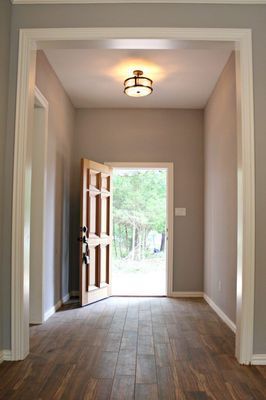 A brightly lit entryway with light gray walls, dark wood plank flooring, a round ceiling light, and an open wooden door.