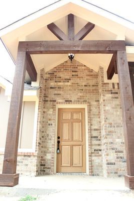 Tan wood front door set in a light brick wall, sheltered by a peaked porch roof with dark brown wooden beams and supports.