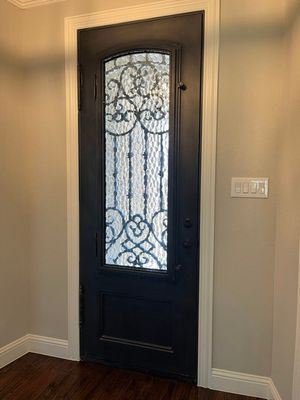 A black front door with a decorative, arched wrought iron glass window panel set in a beige wall with a light switch.