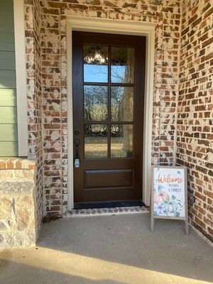 A dark brown wooden door with glass panels sits in a light-colored brick entryway beside a welcome sign on a tripod stand.