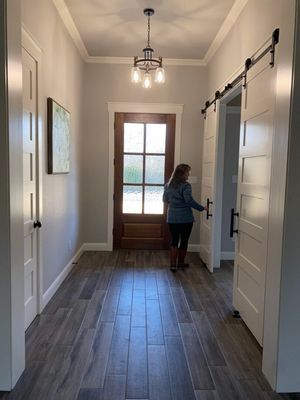 A person stands in a modern hallway with light grey walls, wood-look tile floors, a wooden front door, and white barn doors.