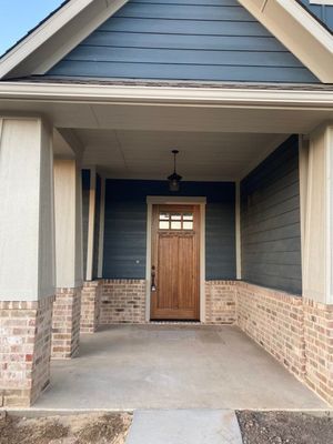 A covered home entrance with a light brown wooden front door, blue lap siding, and a tan brick base.