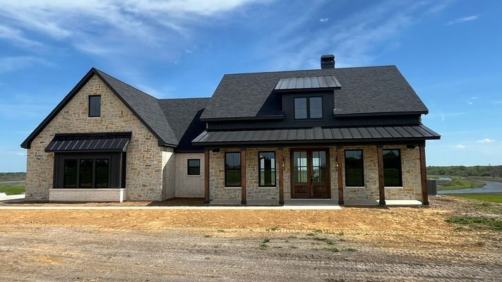 A stone farmhouse with a black metal roof, front porch, and large windows set against a blue sky over a grassy field.