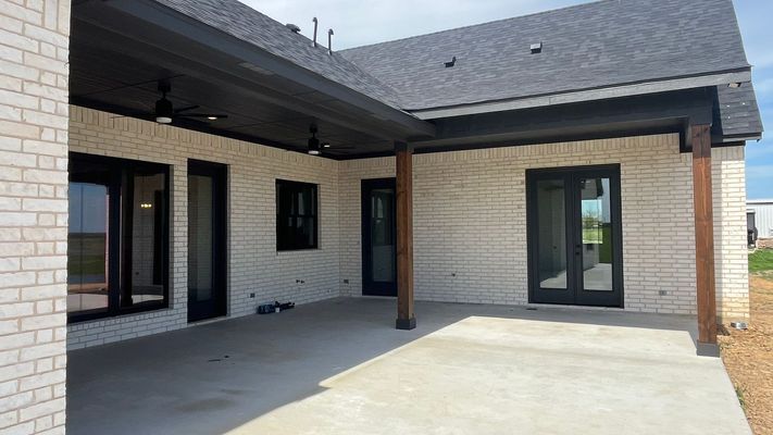 A covered patio with light-colored brick walls, dark trim, black framed windows and doors, and two wooden support posts.