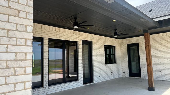 A covered patio with light-colored brick walls, dark trim, a dark wood post, and ceiling fans overlooking a field.