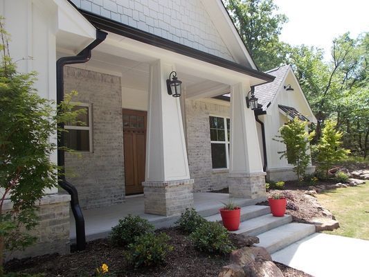 A front porch of a house with stone accents, white pillars, a wooden door, and two red planters on concrete steps.