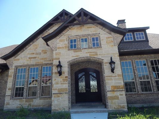 A two-story house exterior featuring tan stone masonry, dark wooden trim, a front double door, and multiple windows.