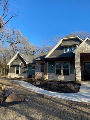 A single-story home with a stone and dark green siding exterior, dark shingled roof, and a concrete walkway in a yard.