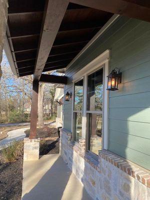 A porch with sage green horizontal siding, stone base, dark wood support beams, and two black outdoor lantern lights.