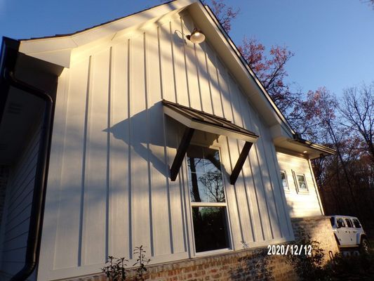 A low-angle view of a white board-and-batten house exterior with a window, small roof awning, and stone base at sunset.