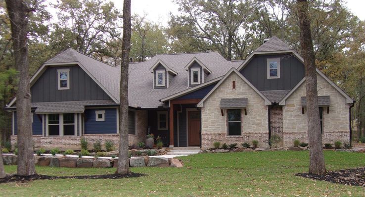 A two-story suburban home with stone and dark blue siding, a shingled roof, and dormer windows, set among mature trees.