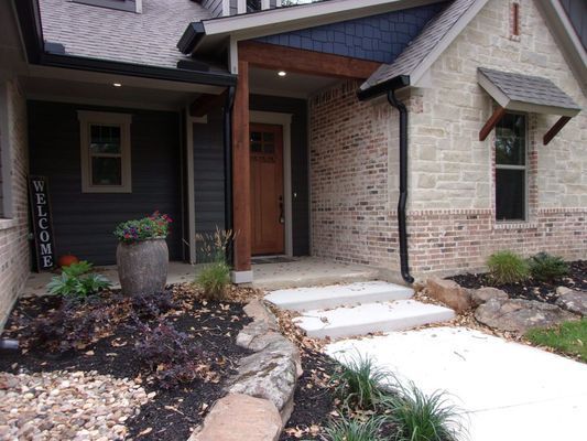A house entrance with a stone wall, wood door, dark grey siding, a potted plant, and a white concrete walkway.