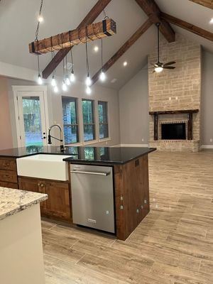 A modern kitchen with a wooden beam pendant light, a dark countertop island, farmhouse sink, and stone fireplace.
