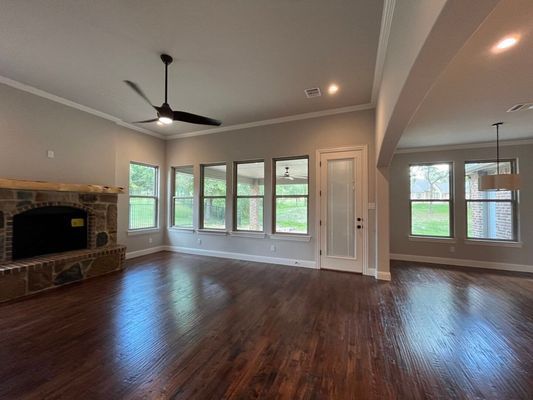 A spacious room featuring hardwood floors, a stone fireplace, large windows, a ceiling fan, and an adjoining open area.