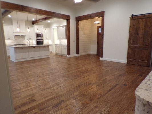 A spacious room with dark wood flooring, open to a white kitchen with a center island and wood-trimmed arched doorways.