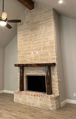 A floor-to-ceiling cream stone fireplace with a thick, dark wood mantel and supports in a room with light wood floors.