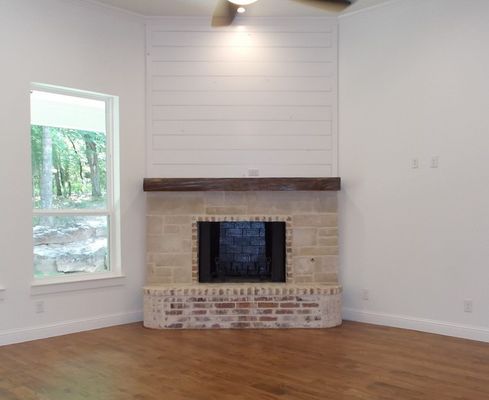 A corner fireplace with stone masonry, a dark wood mantel, and white shiplap walls, beside a window in a bright room.