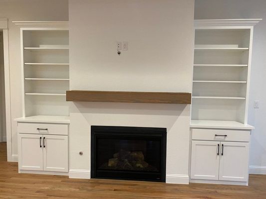A modern white fireplace flanked by symmetrical built-in shelving units with lower cabinets and a wooden mantel.