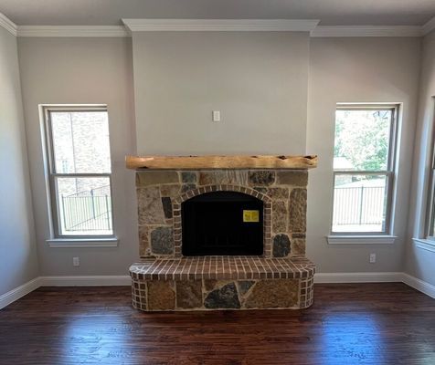 A stone fireplace with a thick wood mantel sits between two tall windows in a room with light grey walls and wood floors.