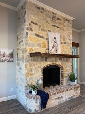 A stone fireplace with a rustic wood mantel, a brick hearth, two potted plants, and wall art in a modern living space.