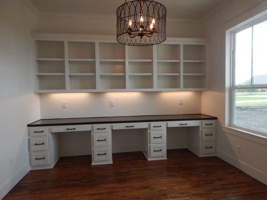 A home office with white built-in shelving and cabinets, a dark wood countertop, and a decorative pendant light.