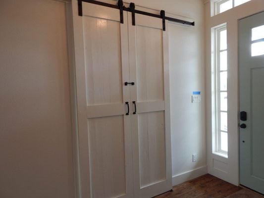 White wooden barn doors on a black sliding rail, installed next to a light blue door with a side window and hardwood floors.