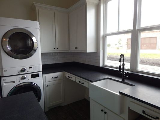 A laundry room with white cabinets, a black countertop, a farmhouse sink, and a stacked washer and dryer unit.