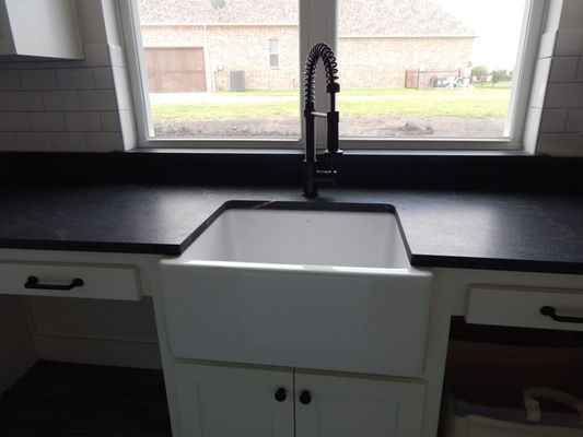 A white farmhouse sink with a tall black faucet, set in white cabinets with a black countertop in front of a window.
