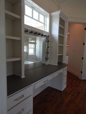 A white built-in vanity station with side shelving, a large mirror with light bulbs, and drawers on a dark wood floor.