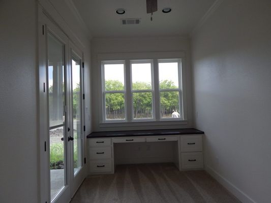 A home office nook with a built-in white desk, dark countertop, and large windows, adjacent to a glass-paneled patio door.