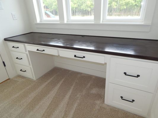 A built-in white desk with a dark wooden countertop and multiple drawers, positioned beneath a large window.