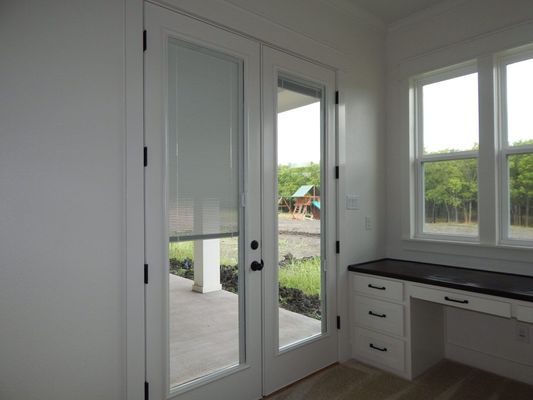 A bright, modern room featuring white French doors with roller shades, white cabinets, and a dark desk surface.