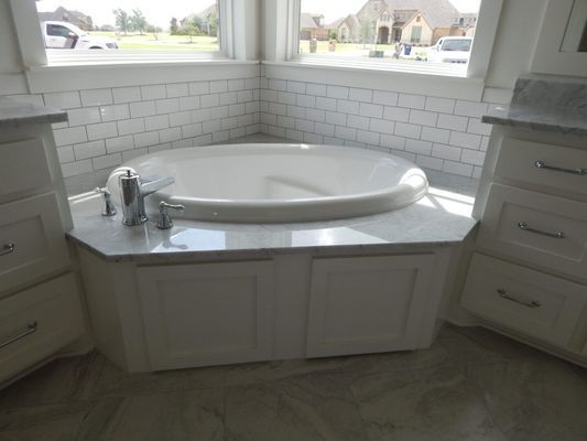 A white oval bathtub set in a marble-topped alcove with white subway tile walls and white cabinetry in a bright bathroom.