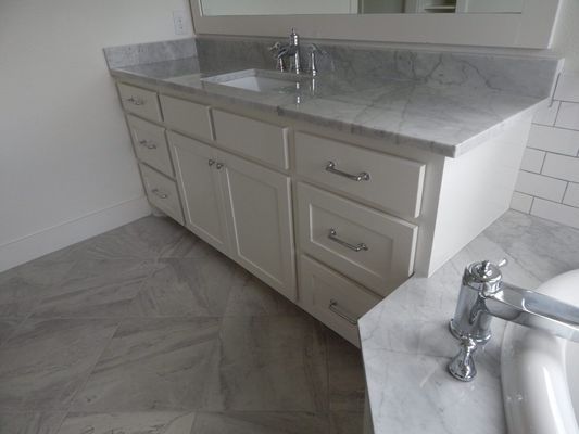 A cream-colored vanity with a marble countertop, silver hardware, and drawers, situated in a white tiled bathroom.