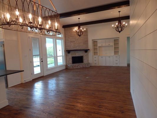 An open-concept living area with dark wood floors, white shiplap walls, a stone fireplace, and rustic chandeliers.