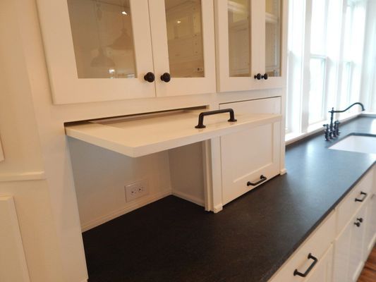 A kitchen counter with white cabinets, including one with an open upward-swinging cabinet door revealing an electrical outlet.