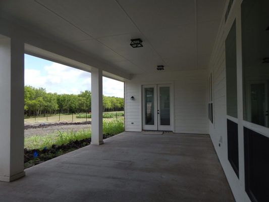 A covered outdoor porch with concrete floors, white pillars, and double glass doors overlooking a field and tree line.