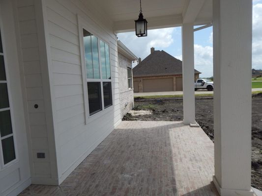 A covered outdoor porch with white walls, a light brick floor, and a hanging pendant light, overlooking a dirt yard.
