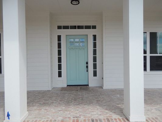 A light blue front door with a window, surrounded by sidelights and a transom, set in a white home porch with brick floors.
