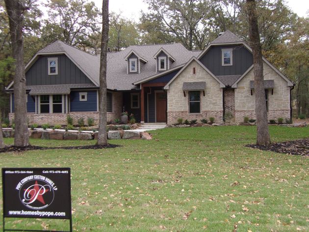 A modern, multi-gabled house with stone and blue siding, set in a wooded area with a real estate sign in the foreground.