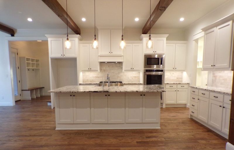 Modern white kitchen with wooden ceiling beams, a central island, and stainless steel appliances over hardwood floors.