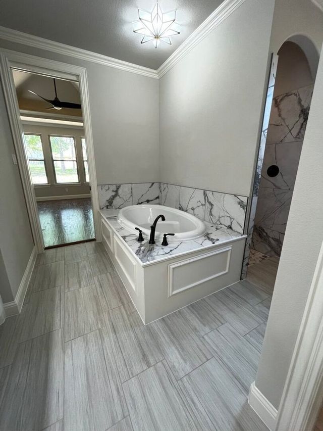A modern bathroom with a white soaking tub set in a marbled deck, grey tiled floors, and a bright, arched entryway.