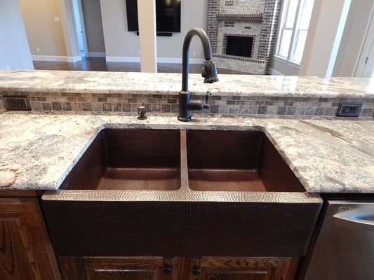 A deep, hammered copper farmhouse sink with a tall dark faucet set in a light granite countertop in a kitchen.