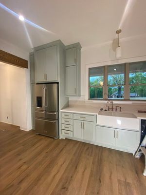 Modern kitchen with light green cabinets, stainless steel refrigerator, white farmhouse sink, and wood-tone flooring.