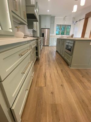 A light sage-green kitchen with light wood flooring, a stainless-steel refrigerator, and a large central island.